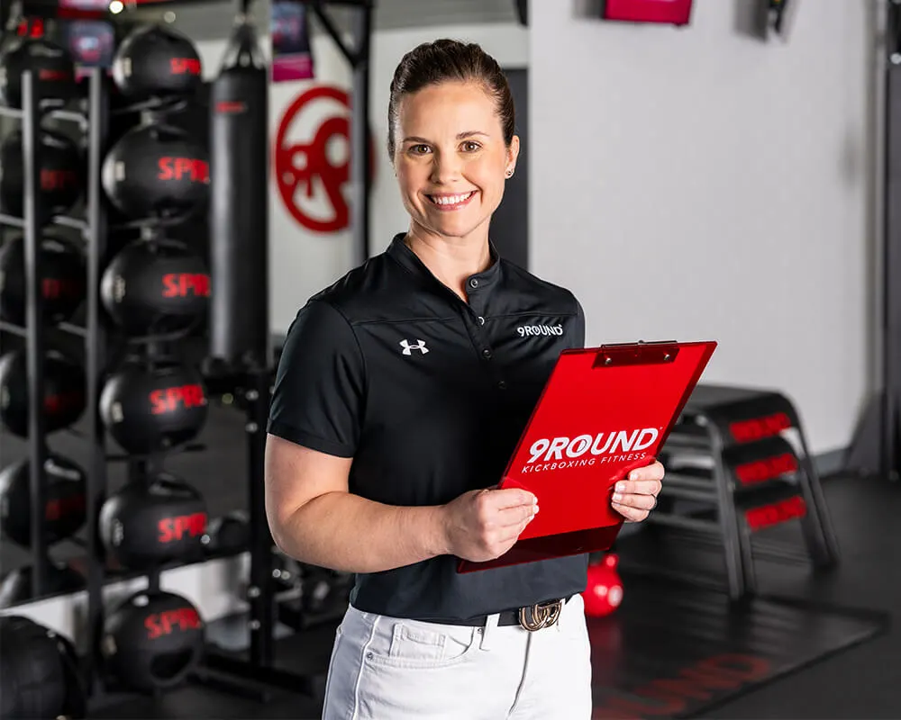 A woman in a black shirt, possibly the franchise owner, holds a red clipboard in a gym filled with equipment, with 9ROUND logos adorning the background.