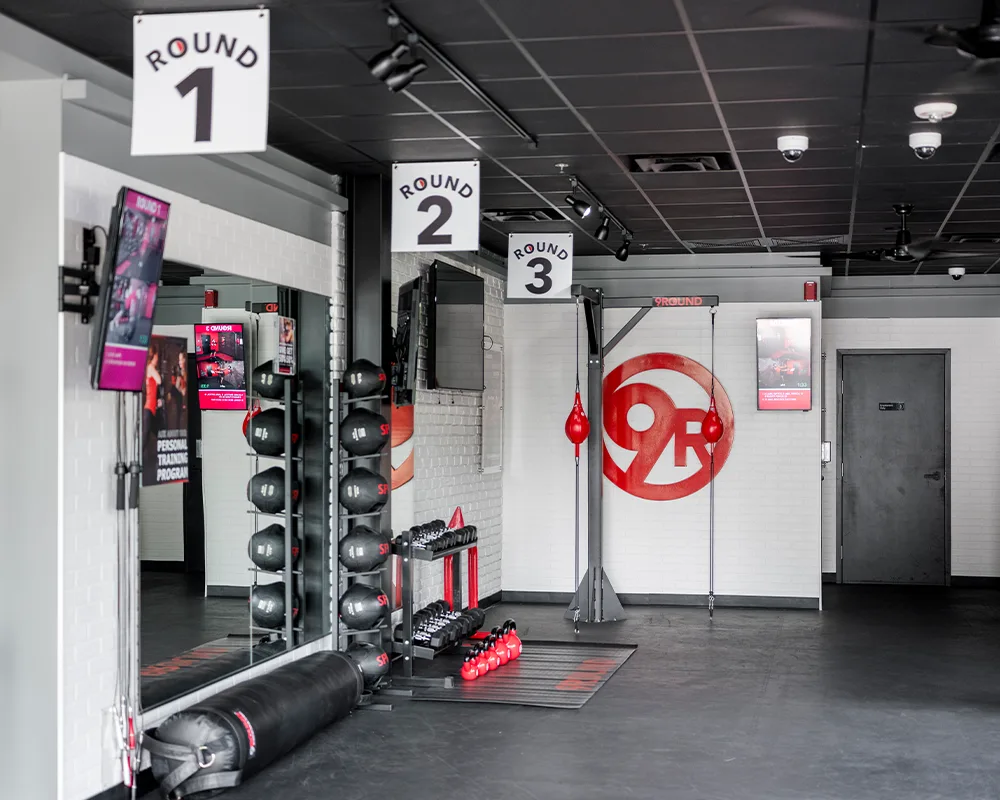 A sleek gym and fitness studio featuring boxing equipment like heavy bags and speed bags. Workout station signs dangle from the ceiling, while weights are neatly stacked on shelves.