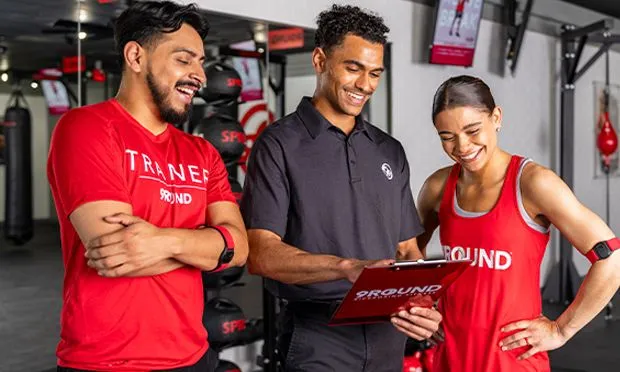 In a professional atmosphere, three people in a gym gather around a clipboard. Two sport red 9ROUND shirts, while the other wears black. Exercise equipment forms the backdrop as they focus intently on their discussion.